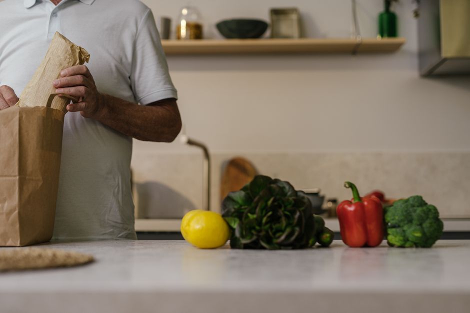 A man unpacks groceries from a paper bag on a kitchen counter with fresh vegetables