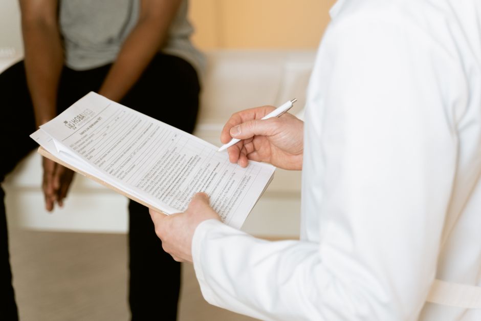 En læge gennemgår en skema på et klemmebræt sammen med en siddende patient