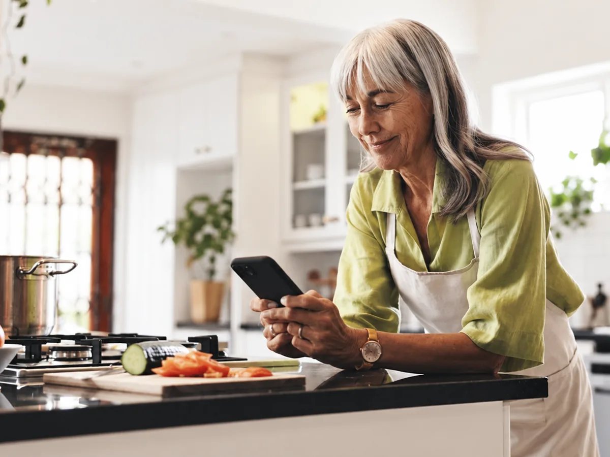 A woman in her kitchen, looking at Kairi on her phone beside the food she is preparing